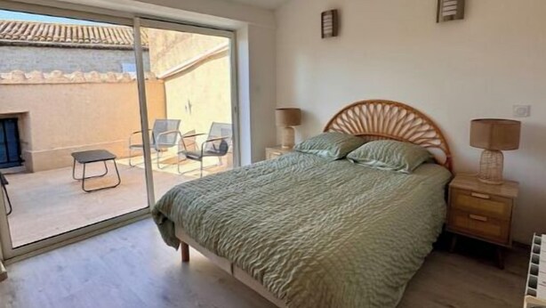 A bedroom with a bed, a nightstand, a lamp, and a sliding glass door leading to an outdoor area with chairs. - La Maison de Sainte-Eulalie (Roubia)