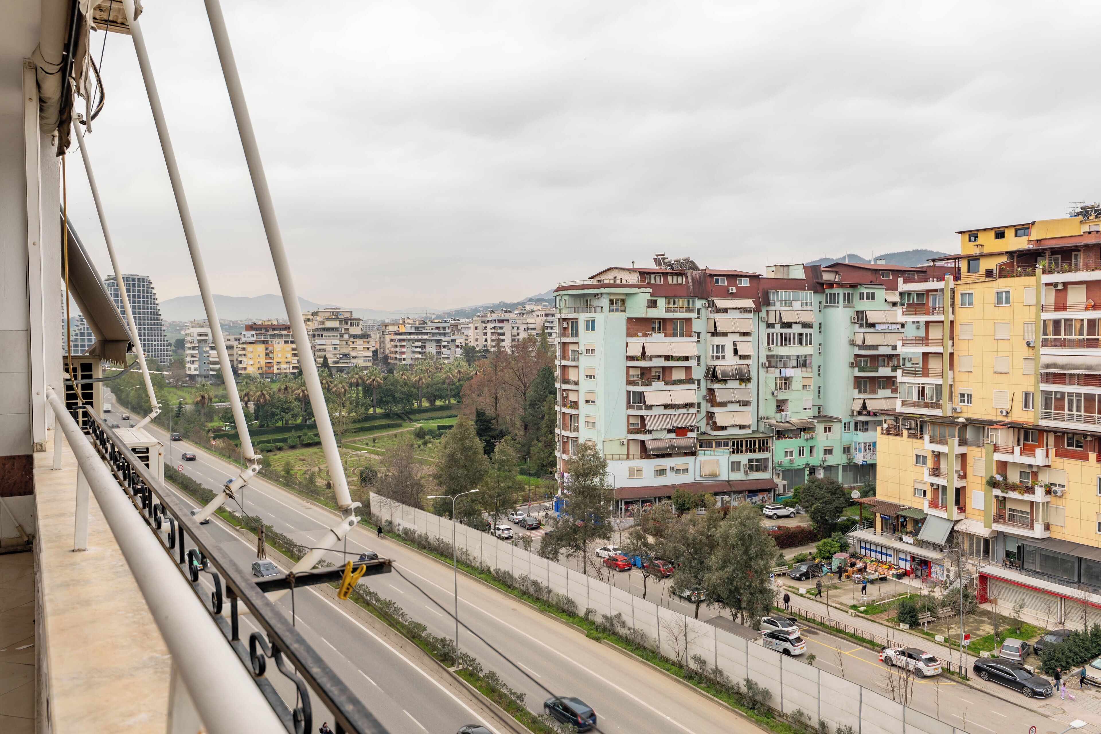 A multi-story building with balconies, a road with cars, and a bridge in the background.