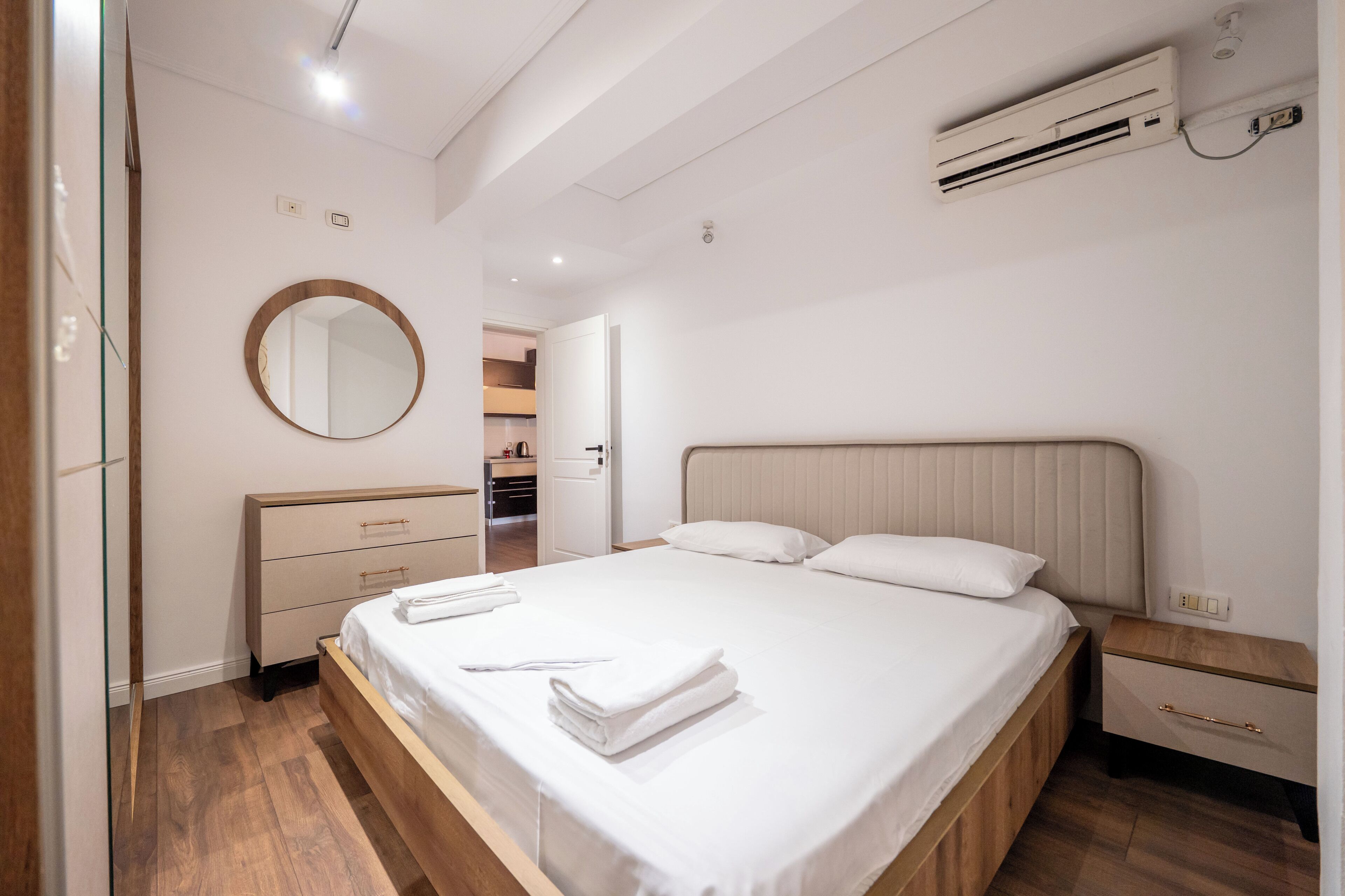 A modern hotel room with a wooden bed, a bedside table, a mirror, and an air conditioning unit.