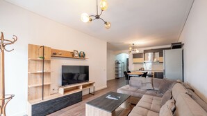 Una sala moderna con un sofá grande, una mesa de centro y un televisor. Se observa una estantería de madera y un área de comedor al fondo.