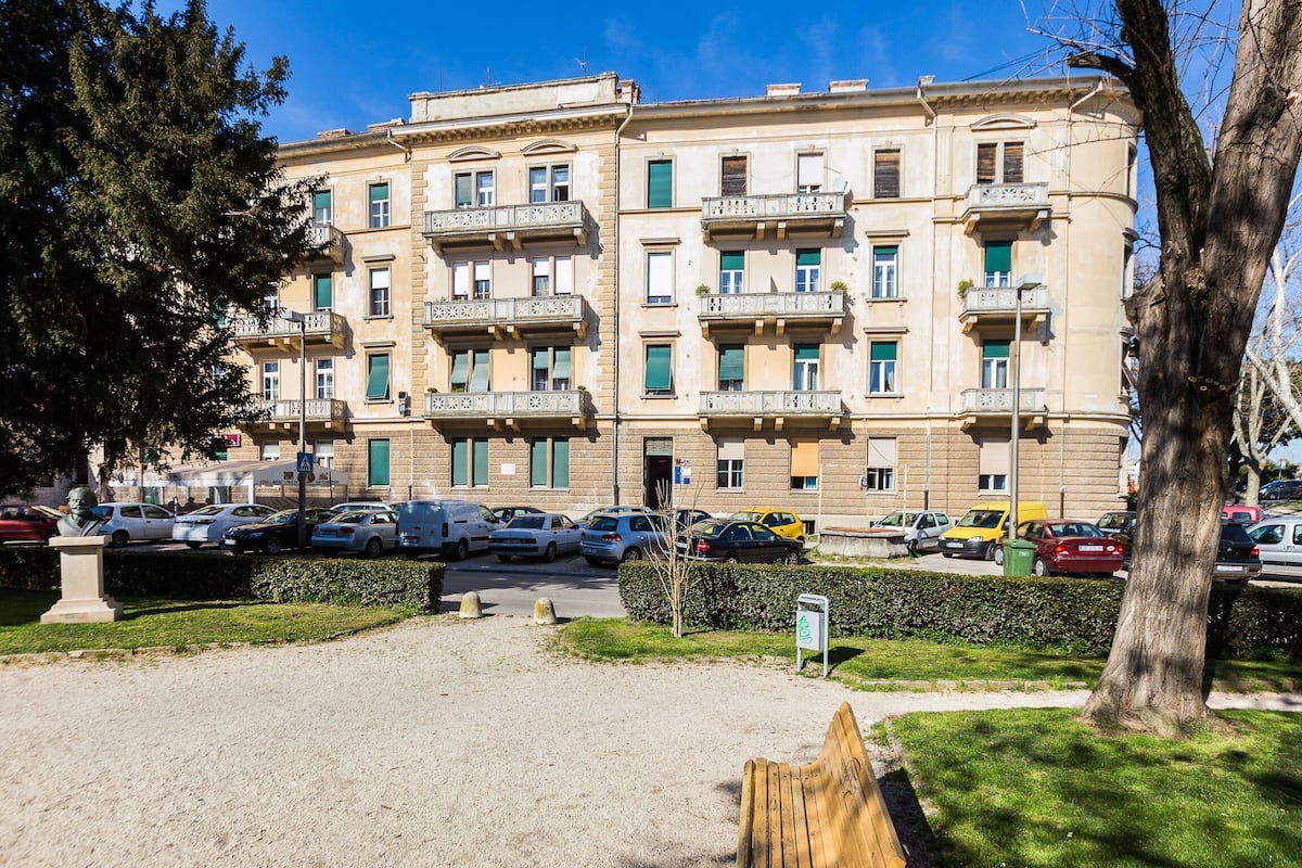 A multi-story building with balconies, a parking lot, and a grassy area with a bench.