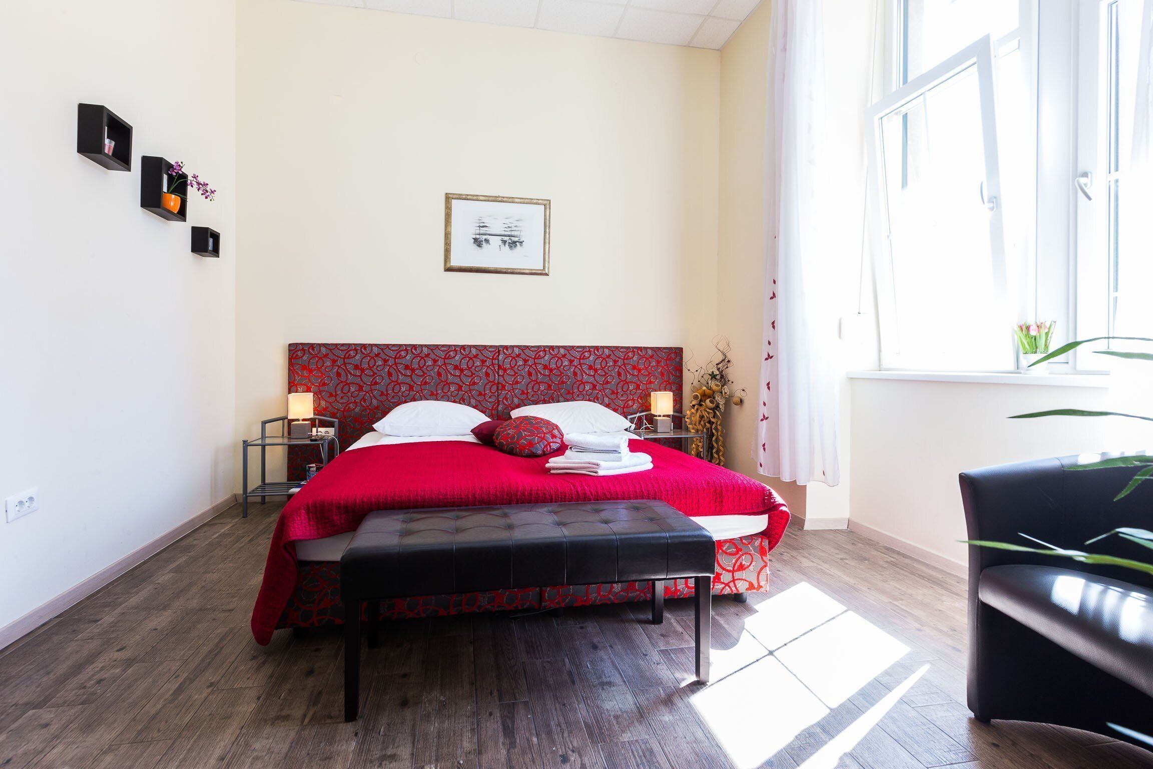 A bedroom with a red patterned bedspread, a black bench, a bedside table with a lamp, and a window with curtains.