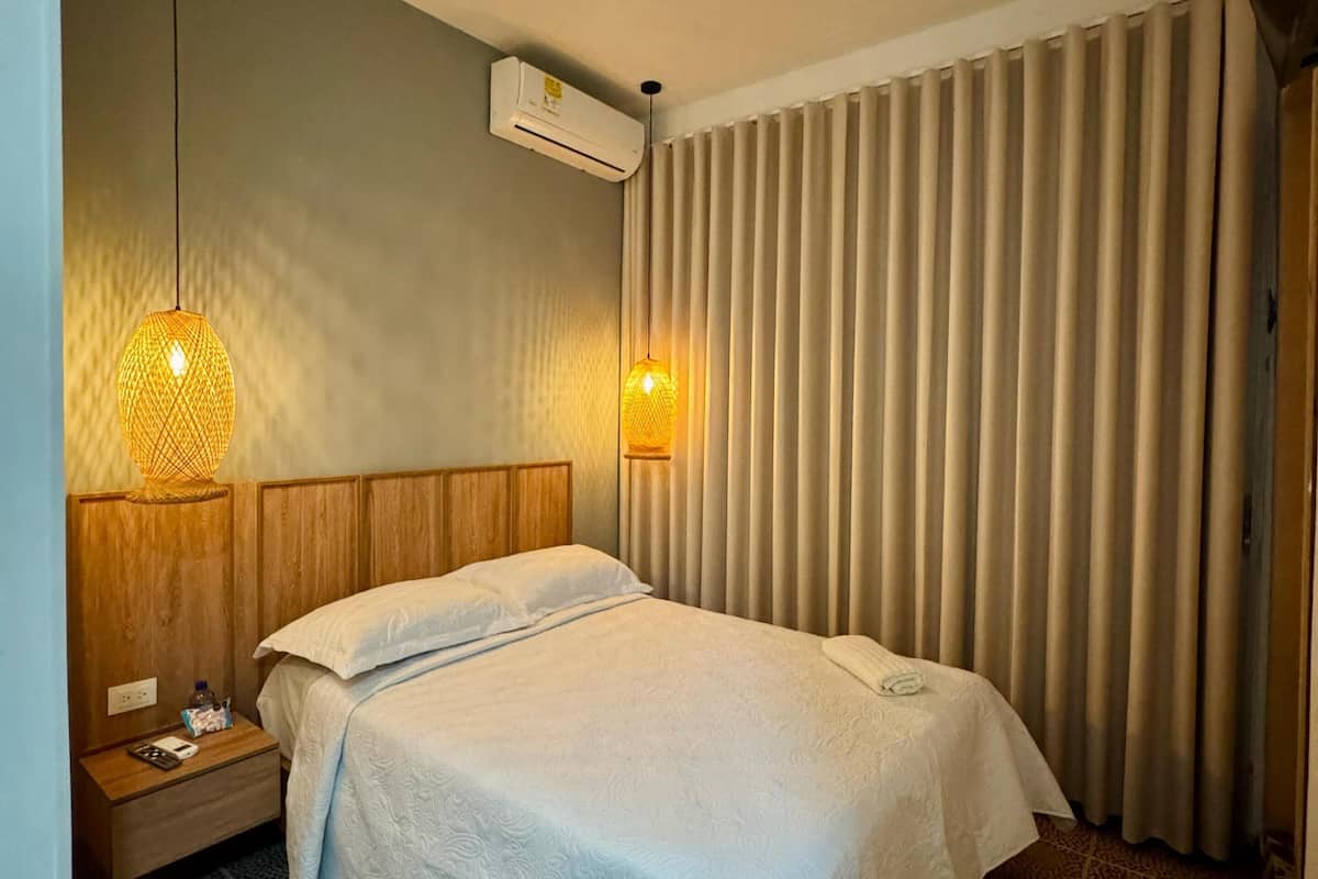 A hotel room with a bed, a nightstand, a wall-mounted air conditioner, and a window with vertical blinds.