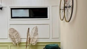A room with a window, a wall-mounted bicycle wheel, a macrame wall hanging, a macrame vase, a green barrel, and a wooden stool. - Treeloft 29 (Girón)