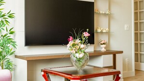 Un salon avec un téléviseur à écran plat fixé au mur, un vase en verre contenant des fleurs posé sur une table basse en bois, et une plante en bambou sur le côté gauche.