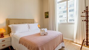 Un lit bien fait, avec du linge de lit blanc et une couverture rose, une lampe de chevet et une fenêtre avec des rideaux.
