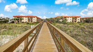A wooden boardwalk leading to a hotel with red roofs and multiple floors, surrounded by greenery and a clear sky.