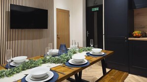 A modern dining area with a long wooden table set for a meal, a flat-screen TV mounted on the wall, and a kitchen area with dark cabinetry and a fruit bowl on the counter.