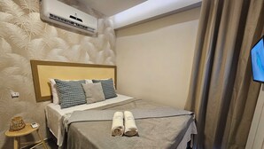 Ein ordentlich bezogenes Bett mit grauer Decke und aufgerollten Handtüchern, ein kleiner Nachttisch mit Korb und eine wandmontierte Klimaanlage.