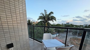 Ein Balkon mit zwei weißen Tischen und Stühlen, einer Glasreling und Blick auf eine Palme sowie Gebäude in der Ferne.