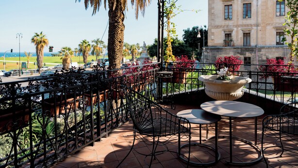 A terrace with a view of the street and a building, featuring a table and chairs, potted plants, and a decorative fountain. - The Prince's "Casena" - Historic home with a sea view (Palermo)