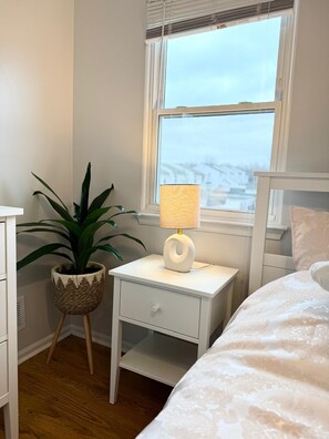 Een slaapkamer met een bed, een nachtkastje met een lamp, een potplant en een raam met zonwering.
