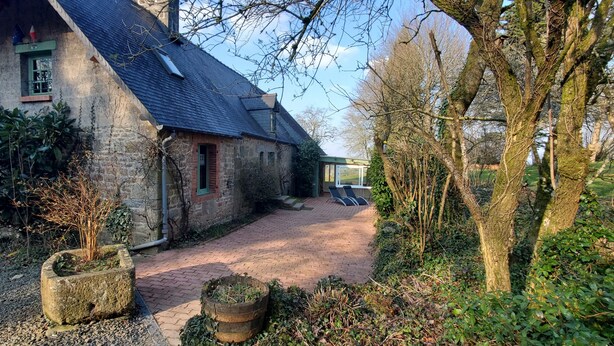 Une maison en pierre avec un toit en pente, un chemin en briques menant à un espace aménagé pour s’asseoir, et une végétation luxuriante tout autour.
