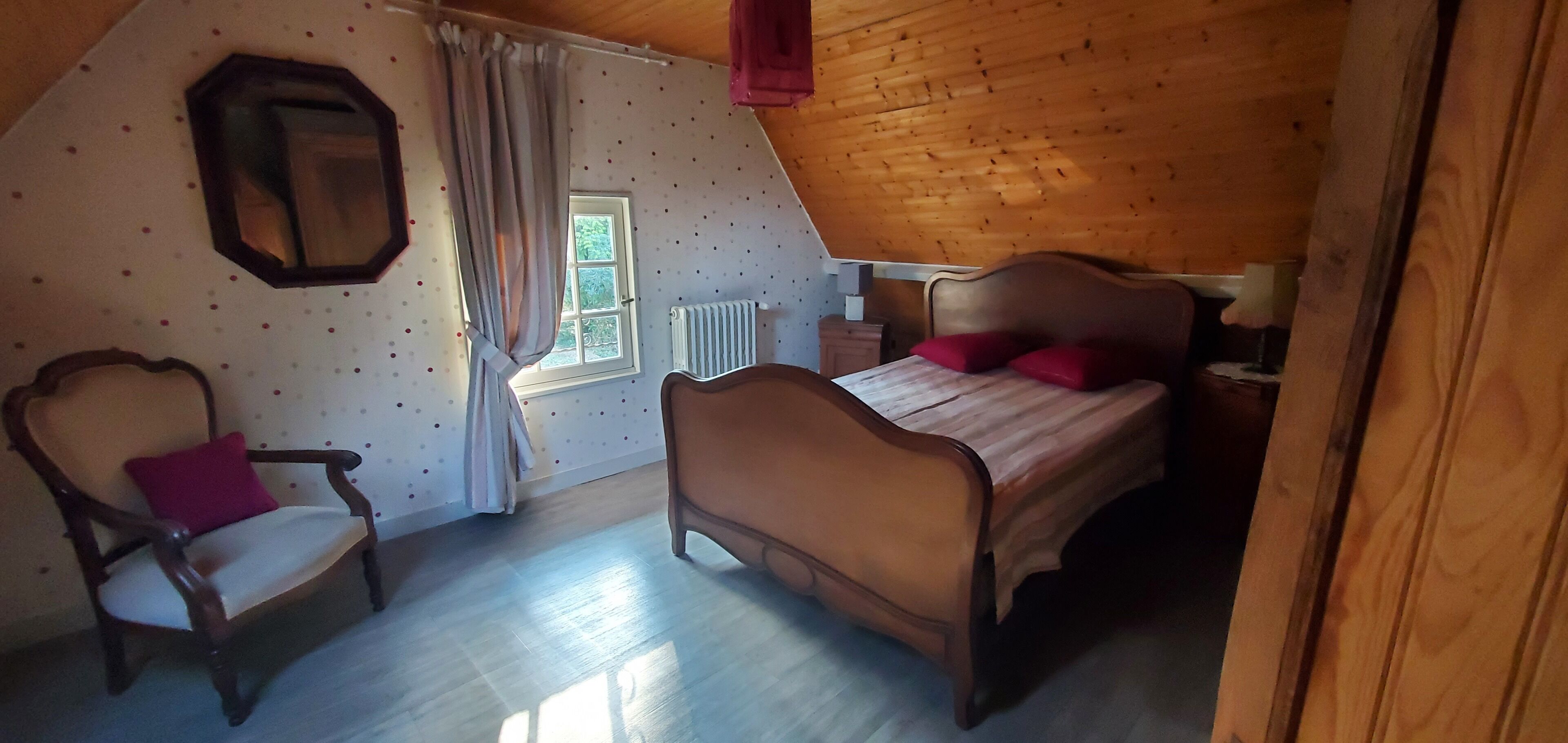 Una camera da letto con soffitto in legno, un letto con cuscini rossi, una sedia con cuscino rosso, una finestra con tende e un termosifone.