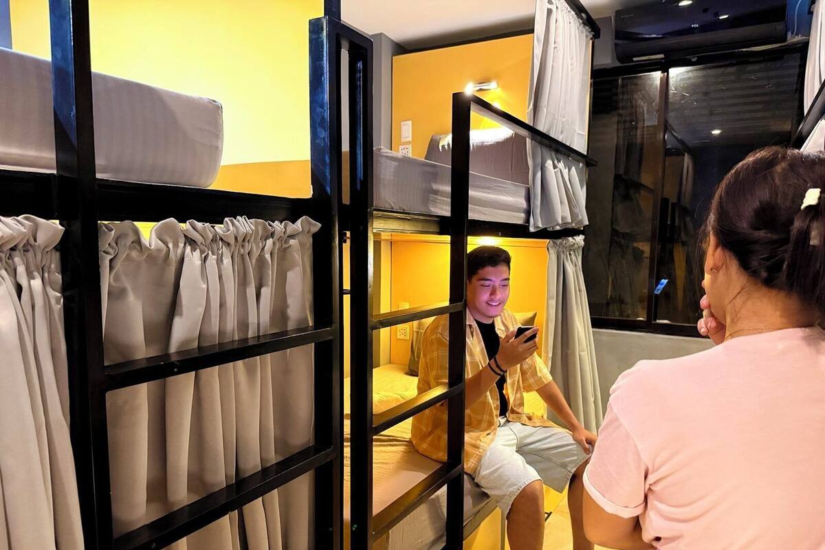 A bunk bed room with a yellow wall, a window with curtains, and a person using a smartphone.