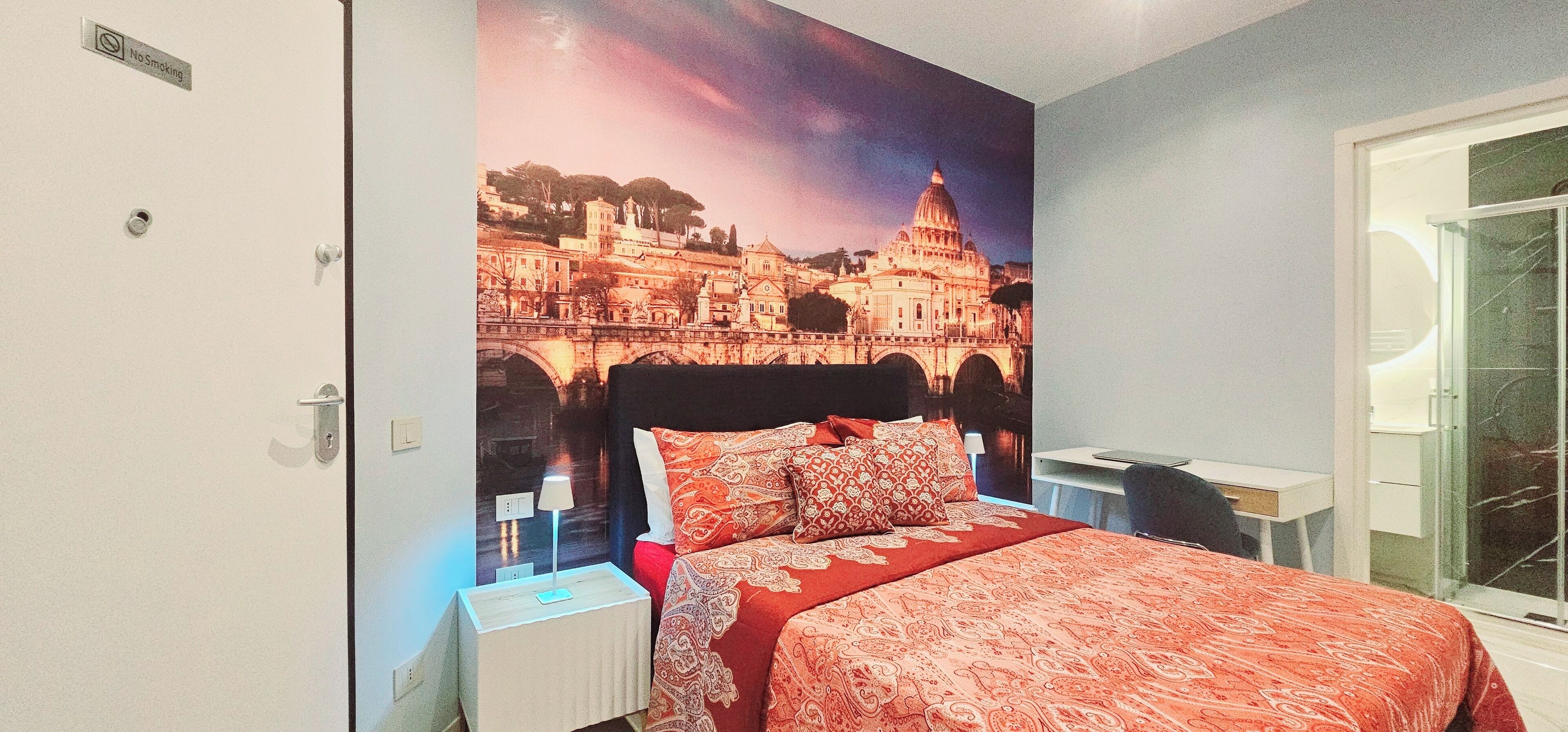 Ein Hotelzimmer mit einer großen Stadtansicht als Wandgemälde, einem Bett mit rot gemustertem Überwurf, einem Nachttisch mit Lampe sowie einem kleinen Tisch mit Stuhl.