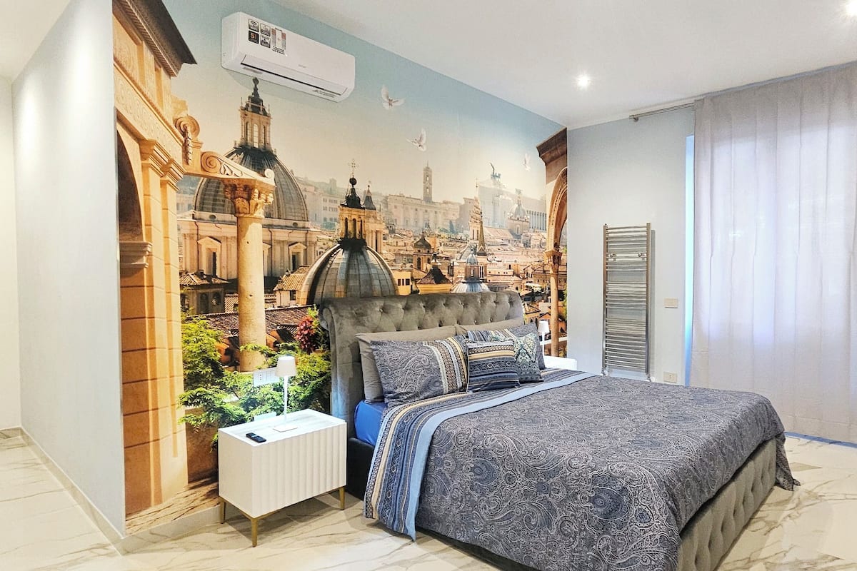 A bedroom with a large bed, a nightstand, a radiator, and a wall mural of a cityscape.