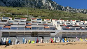 Un hotel frente al mar con varios balcones con vistas a la playa, una fila de kayaks de colores y un telón de montañas al fondo.