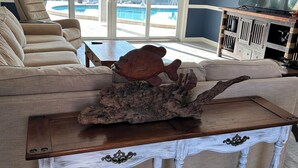 Ein Wohnzimmer mit einem beigefarbenen Sofa, einem Holztisch mit einer Fischskulptur, einem Deckenventilator und einem Fernseher auf einer Holzkommode.