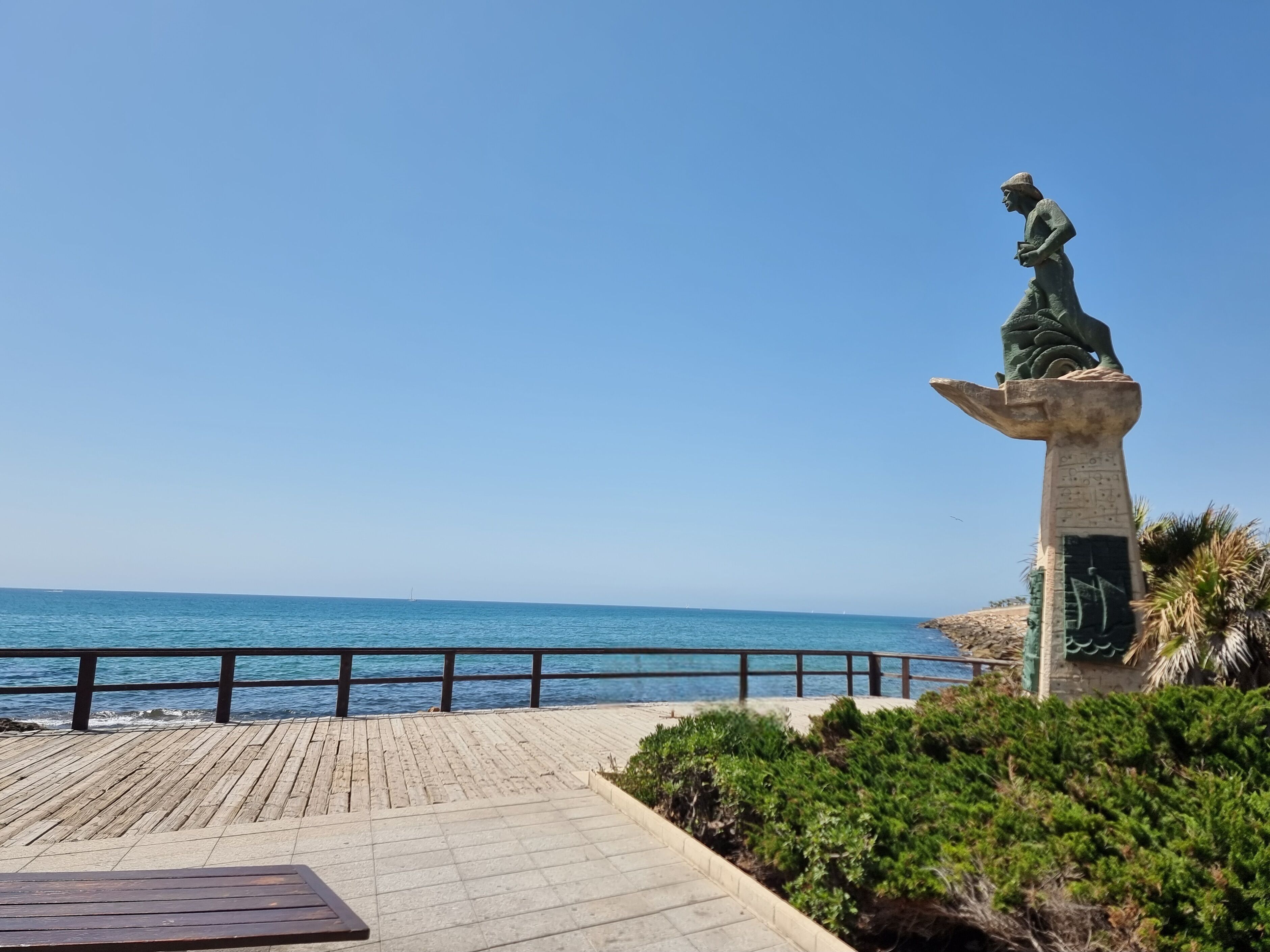 Une statue sur un piédestal, au bord de la mer, une promenade en bois et une rambarde.