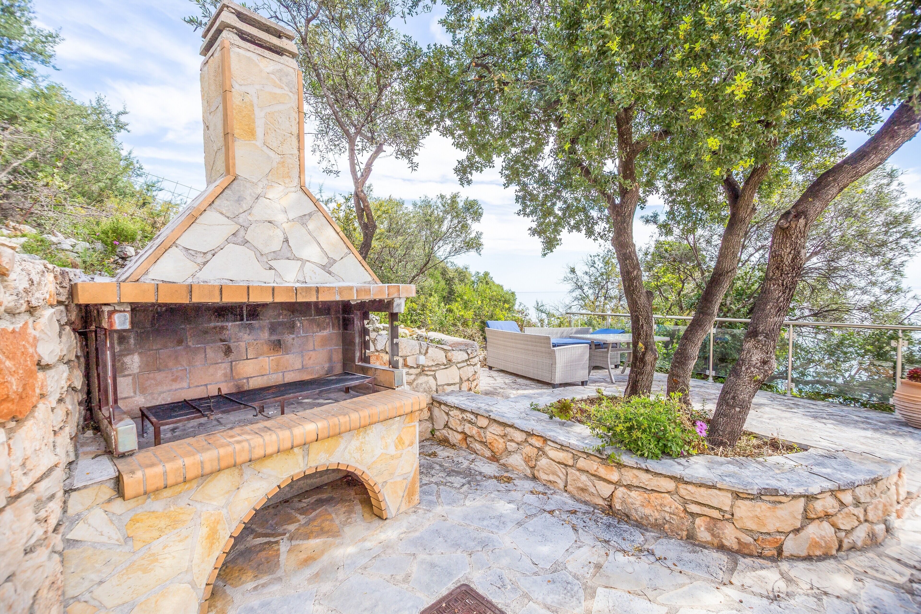An outdoor stone fireplace with a chimney, surrounded by stone walls and greenery.