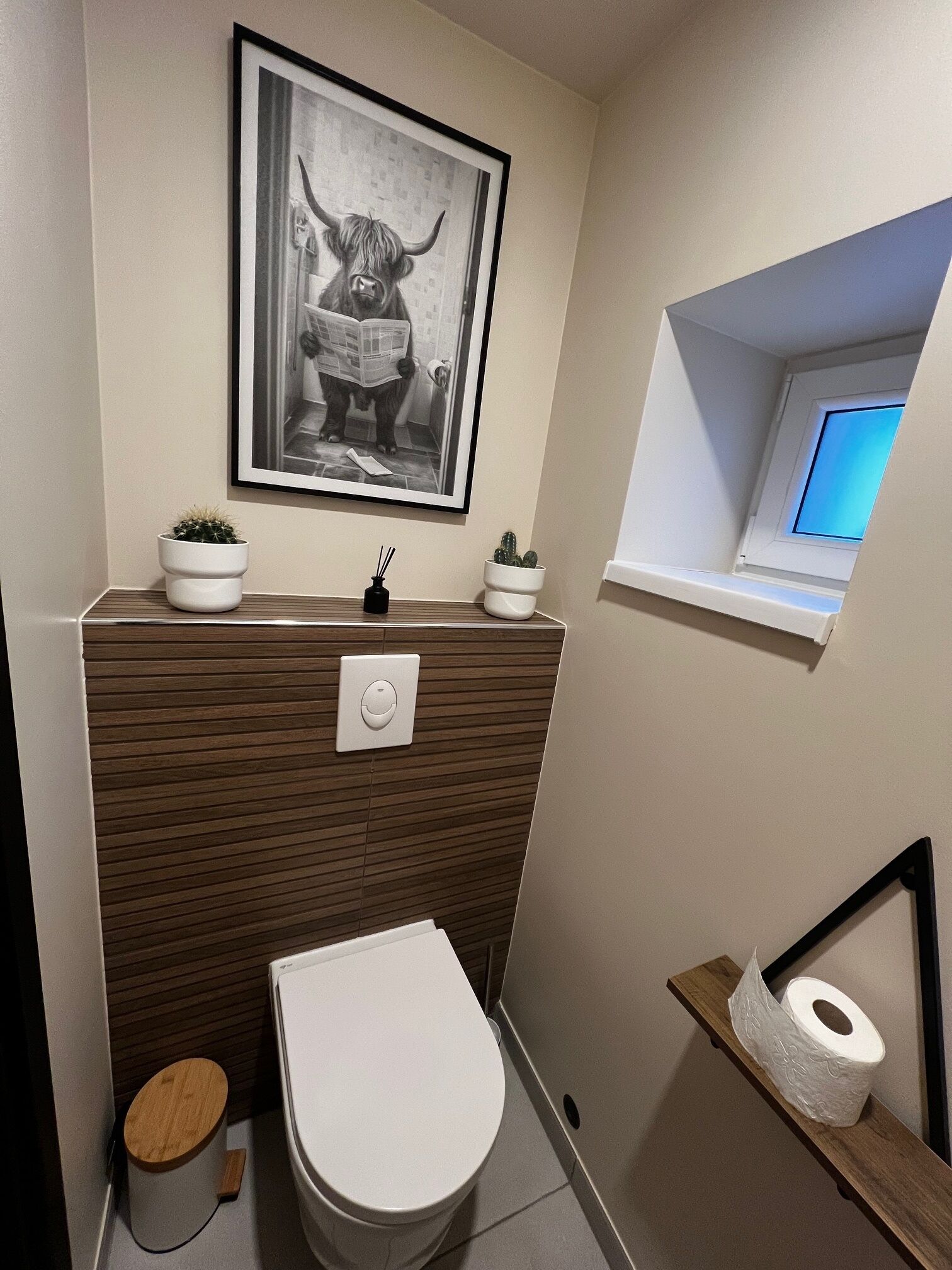 A compact bathroom with a toilet, a sink with two potted plants, a framed picture above the sink, a small window, and a wooden shelf with toilet paper.