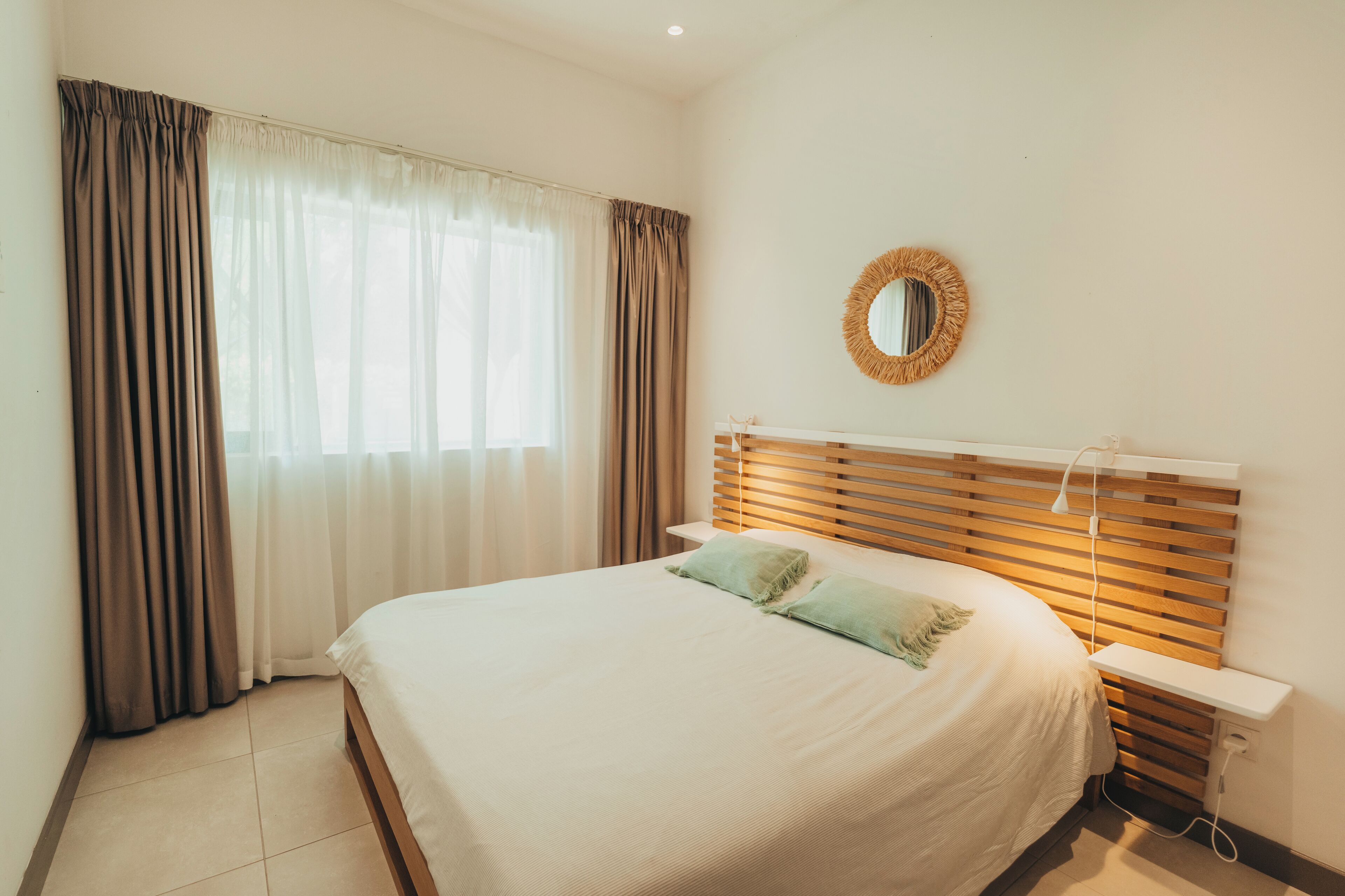 A neatly made bed with a wooden headboard, a bedside table, and a round mirror on the wall.