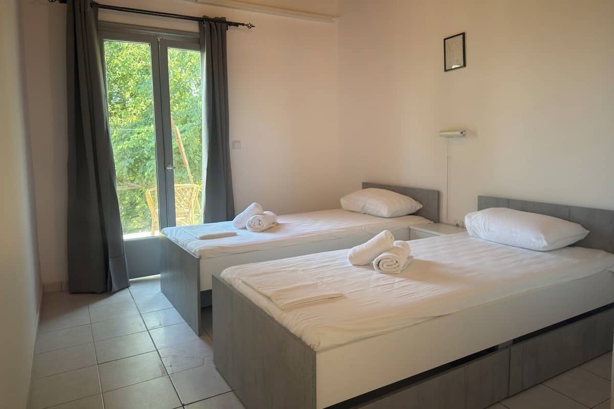 A hotel room with two beds, a window with curtains, a wall-mounted air conditioner, and a small picture frame.