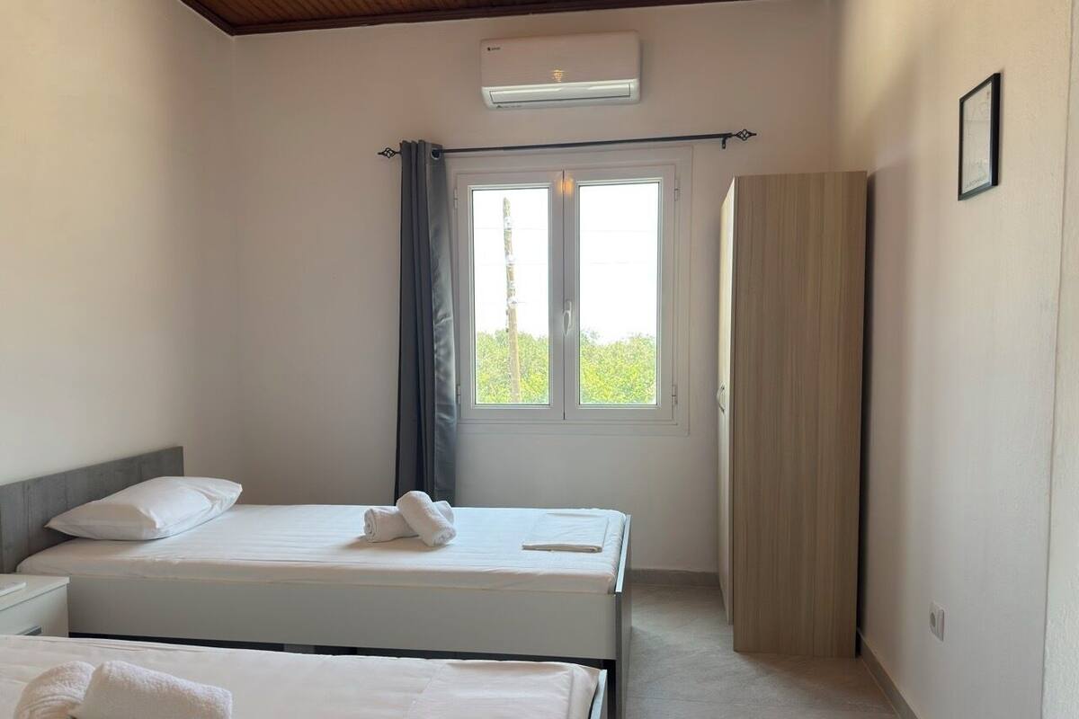A hotel room with two beds, a window with curtains, a wooden door, and a wall-mounted air conditioner.