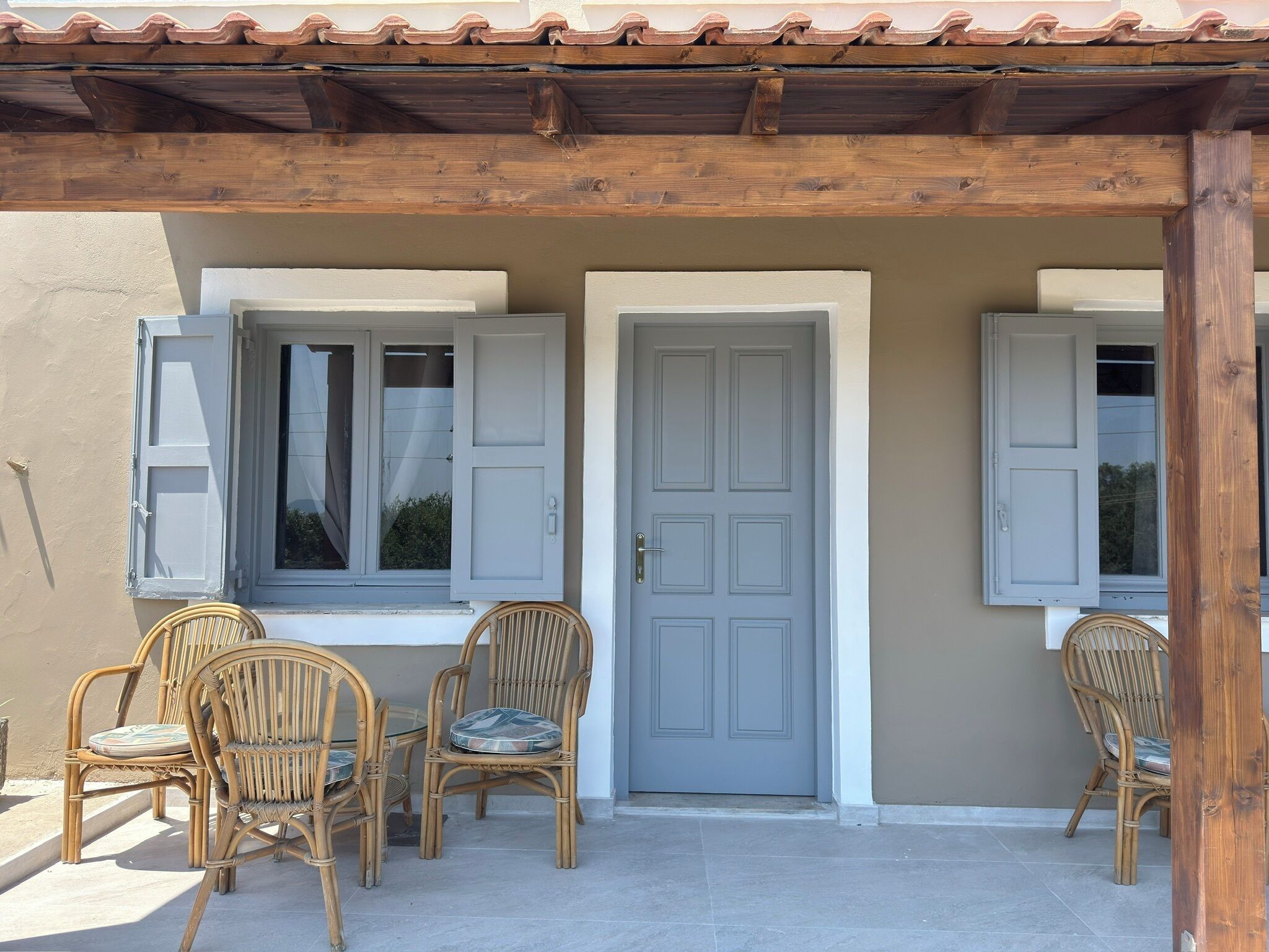 A covered patio with two wicker chairs and a table, a grey door, and windows with shutters.