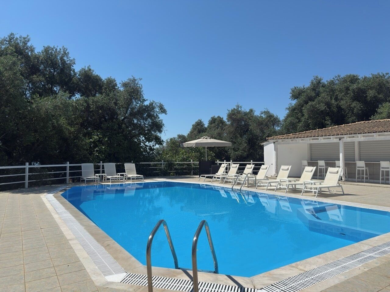A rectangular swimming pool with clear blue water, surrounded by a tiled deck. There are white lounge chairs and a white umbrella on the deck.