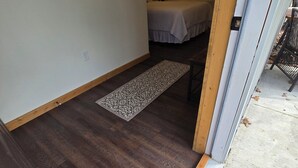 Una habitación de hotel con suelo de madera, una cama con colcha blanca, un espejo redondo pequeño en la pared y un cuadro enmarcado.