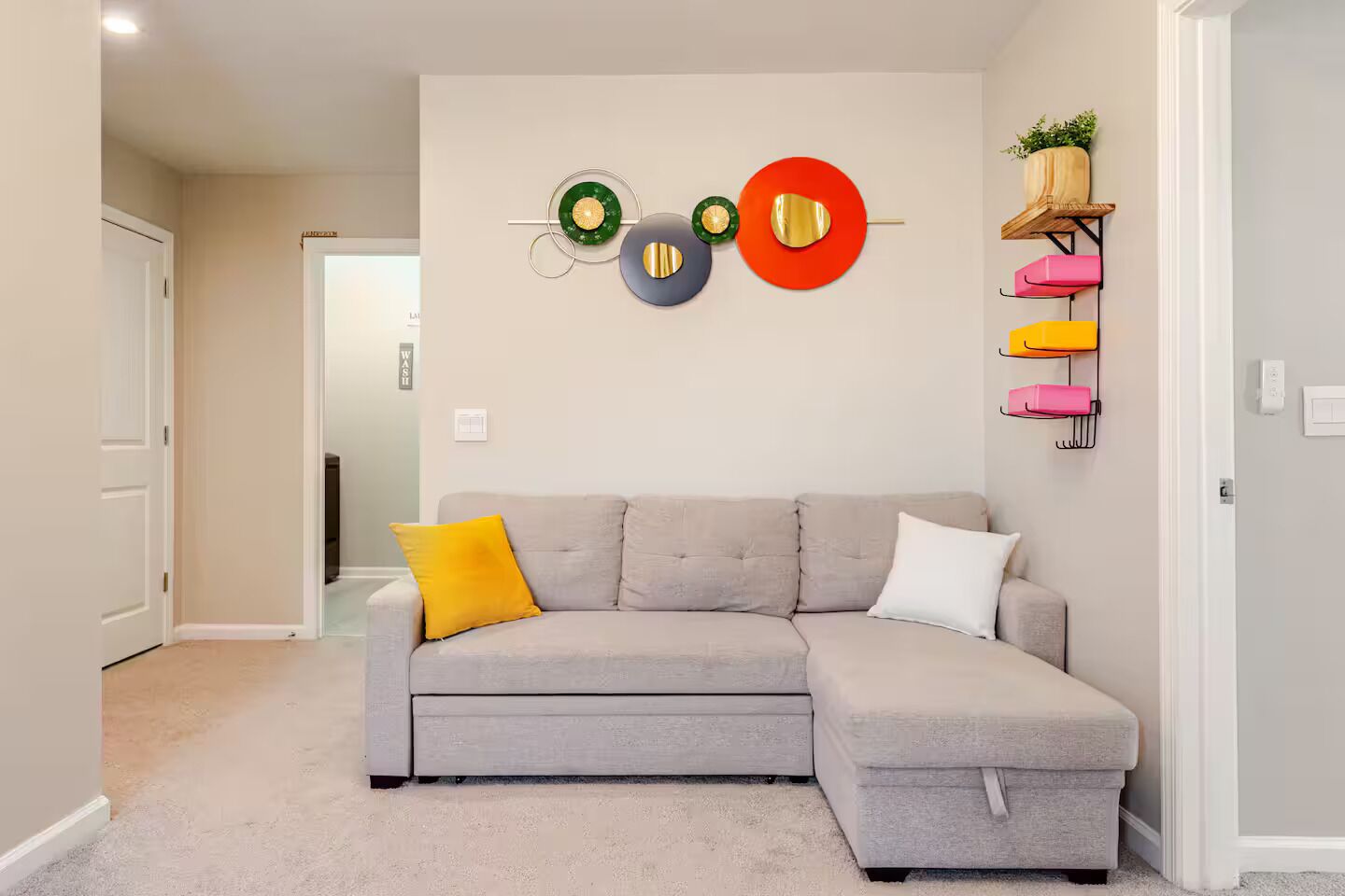 A living room with a grey sectional sofa, a yellow pillow, and a red and green wall decoration. There is a shelf with colorful storage boxes and a potted plant.