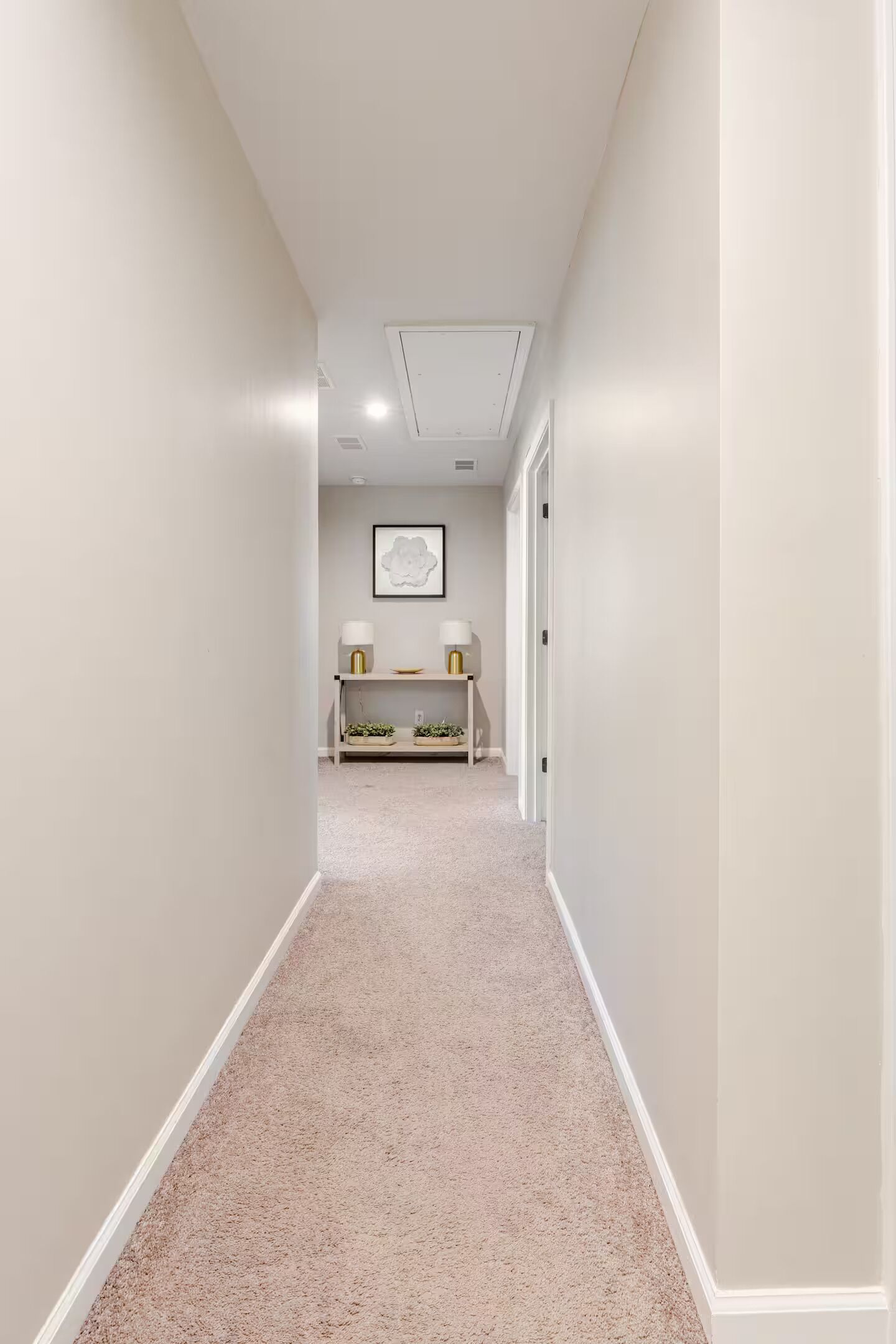 A narrow hallway with a carpeted floor, a white door at the end, and a small seating area with a lamp and a framed picture on the wall.