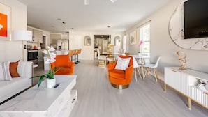 Ein modernes Wohnzimmer mit einer Couch, orangefarbenen Sesseln, einem Essbereich und einer Küche im Hintergrund.