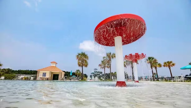 Ein Wasserbrunnen mit roter Oberseite und weißem Unterteil, umgeben von Palmen und einem Gebäude mit rotem Dach.