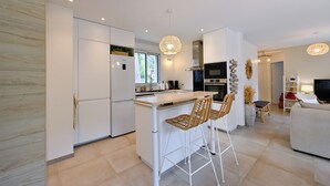 A modern kitchen with white cabinetry, a central island with bar stools, and a dining area with a sofa and TV.