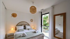 A bedroom with a large bed, bedside lamps, a wicker ceiling light, a round mirror, and a view of greenery outside.