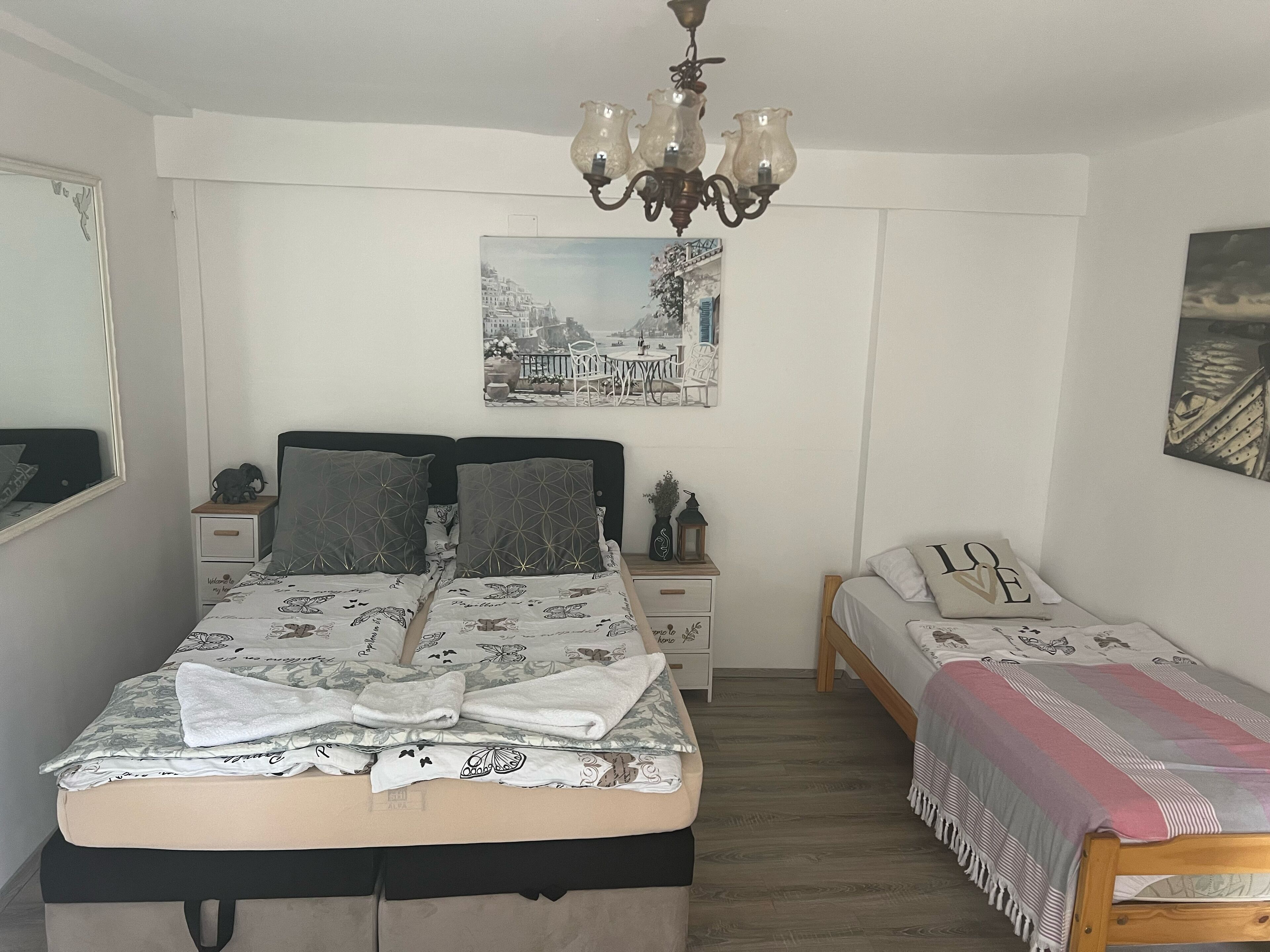 A bedroom with a large bed, a smaller bed, a nightstand, a dresser, a mirror, a chandelier, and a cityscape poster on the wall.