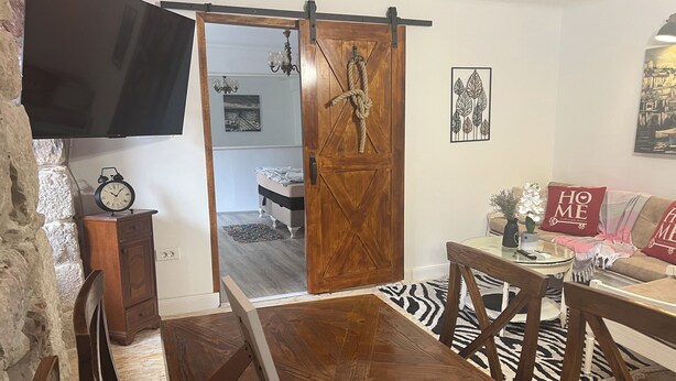 A dining area with a wooden table and chairs, a sliding barn door, a television mounted on a stone wall, and a sofa with decorative pillows. - Dora (Bakar)