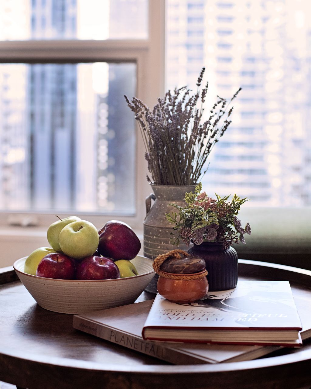 A table with a bowl of apples and pears, a vase with dried flowers, a small pot with a plant, and a book titled "PLANETFALL".