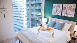 Ein Hotelzimmer mit einem großen Bett, einem Nachttisch mit Lampe, einem Tablett mit Frühstücksangeboten und Blick auf die Stadtlandschaft durch ein großes Fenster.