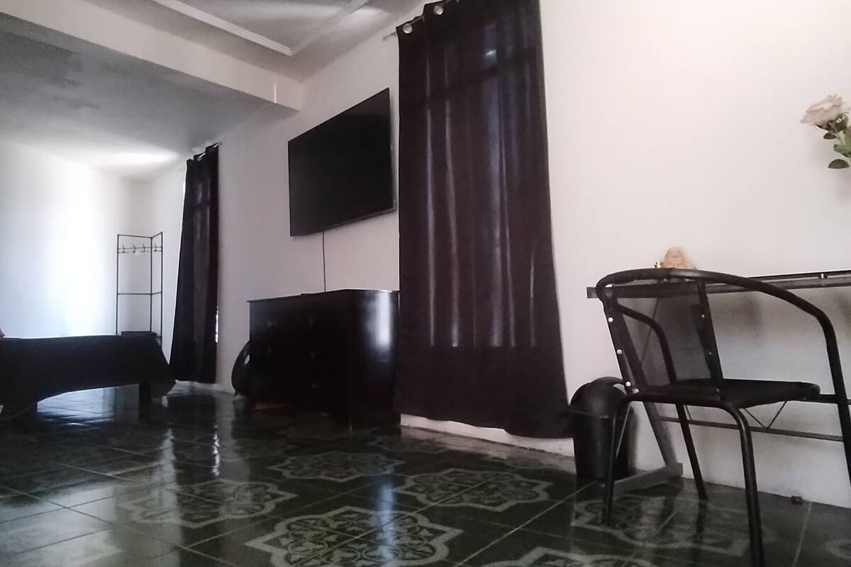 Una habitación con un banco, un televisor montado en la pared, una mesa con sillas y un jarrón con flores.