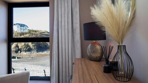 A room with a view of a beach, a wooden table with a lamp and decorative items, and a window with curtains. - Graftons, Little Haven (Little Haven)