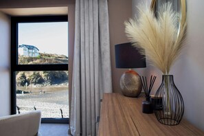 Quarto com vista para a praia, mesa de madeira com abajur e objetos decorativos, e janela com cortinas.