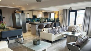 A modern living room with a sofa, coffee table, and chairs. There is a kitchen area with a refrigerator and a dining table. - Graftons, Little Haven (Little Haven)