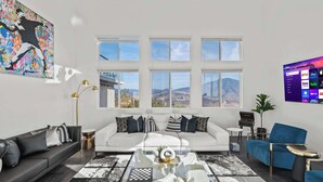 A modern living room with a white sofa, a black sofa, a glass coffee table, and a blue armchair. There are multiple windows providing a view of the mountains. - 5000sf Luxury 5BR, Hot Tub, Panoramic Views, Games, Fire Pit (Spring Valley)