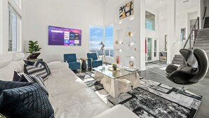 A modern living room with a glass coffee table, white sofas, blue chairs, and a large TV displaying streaming services. - 5000sf Luxury 5BR, Hot Tub, Panoramic Views, Games, Fire Pit (Spring Valley)