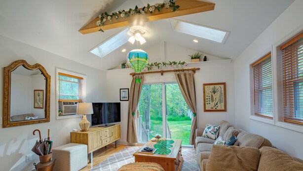 A spacious living room with a sofa, a coffee table, and a television. There is a window with curtains, a ceiling fan, and a decorative hanging light. - The Vibrary Inn, a Fantasy Minnewaska Escape. Minutes from Hikes and Waterfalls. (Kerhonkson)