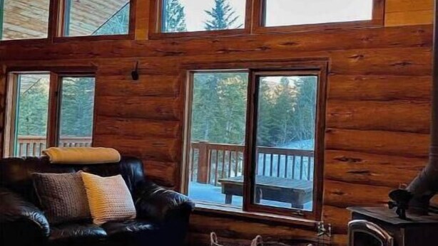 A log cabin interior with a fireplace, leather furniture, and large windows offering views of the outdoors. - Stunning Custom Made Log Home- on 30 acres (Grand Forks)